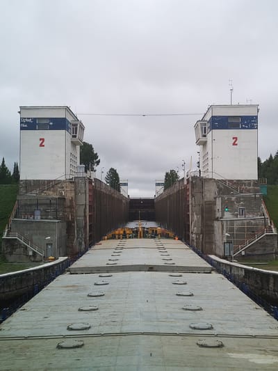 The image shows a canal lock in Анхимово, Russia, specifically Lock No. 2 on the Volga-Baltic Waterway. The photograph is taken from the deck of a barge or boat inside the lock. The lock is flanked by two identical control towers, each painted white with a blue band and marked with a red "2". The towers have multiple windows and antennae on top. The lock walls are made of concrete and steel. A series of concrete slabs with access hatches covers the deck of the barge. Beyond the lock gates, a yellow piece of machinery is visible. The sky is overcast and gray. The sides of the canal are lined with grass and trees.