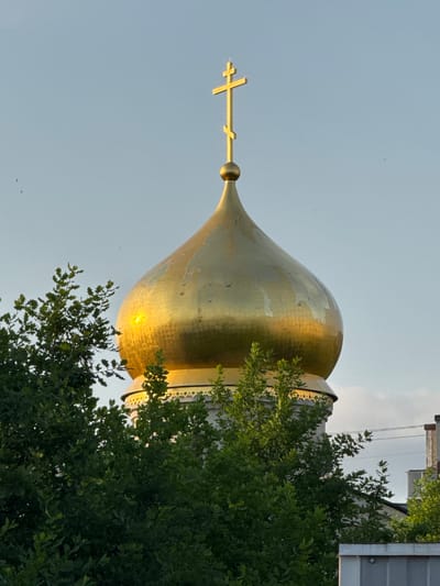 An Orthodox Christian church dome, made of gold-plated material, is visible against a pale blue sky. The dome is topped with a golden Orthodox cross. Lush green trees partially obscure the view of the dome and the church structure below, which appears to be made of white stone with decorative patterns. The scene is captured during daylight, with sunlight reflecting brightly off the dome. The setting suggests an outdoor environment, possibly in the city of Kirishi, Russia, where such religious architecture is common. There are no people or animals visible in the image.