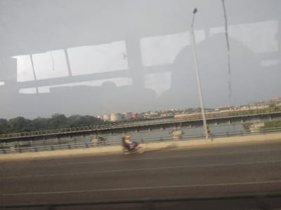 A view from inside a vehicle captures a bridge stretching over water, lined with trees and distant buildings. Storage tanks are visible in the background. A motorcyclist travels along the road on the foreground bridge. Reflections are present in the window, including shadows of people and the vehicle's interior. A streetlight stands tall beside the bridge.