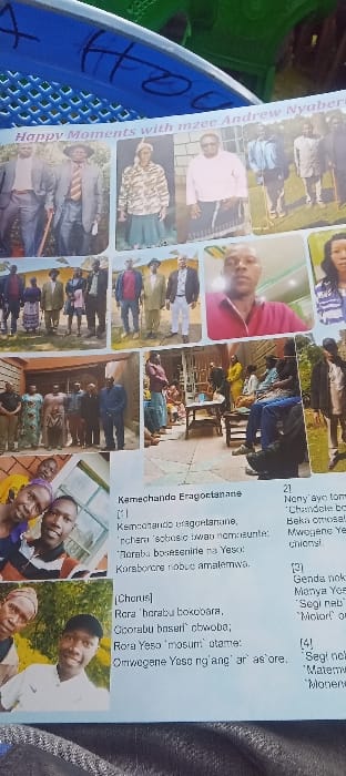 The image displays a printed page, likely from a program or commemorative booklet, resting on a blue, perforated plastic surface that bears black handwritten markings. The page features a collage of color photographs and text, with the prominent horizontal title "Happy Moments with mzee Andrew Nyaberi" across the top.
The photographs depict various individuals and groups. These include multiple outdoor group portraits of men and women in both formal and casual attire, some holding walking sticks. Individual portraits show men and women, one notable selfie of a man in a red polo shirt and other informal selfies with younger subjects. A large central photograph captures an indoor or covered outdoor gathering where several people are seated on benches around a low table, while others stand, with one person in a maroon top central to the scene on a tiled floor. Other photos show individuals walking or posing outdoors.
Below the photographs, a significant portion of text is visible, appearing to be song lyrics. The text begins with "Kamechando Eragoctanane" and includes a chorus section, written in a language other than English, likely a local Kenyan language. The immediate environment of the printed page includes the textured blue surface and a blurred green object in the background. The specific location in Esise ward, Kenya, is not discernible from the visual or textual content of the image.
