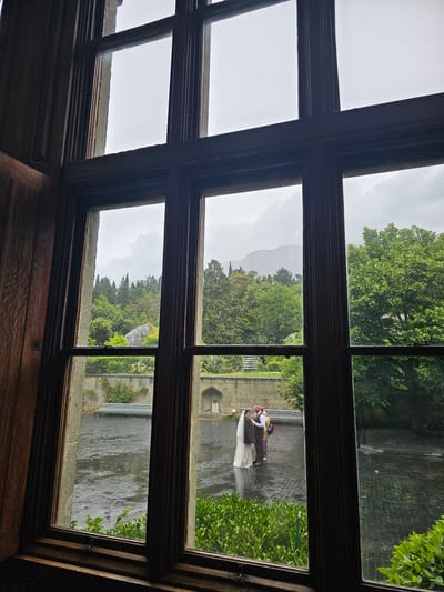 View from a window in Алупкинский городской совет, Ukraine, showing a couple standing on a stone bridge in what appears to be a park or garden. The trees and sky are overcast, suggesting a rainy day. The architectural style visible through the window includes traditional woodwork.