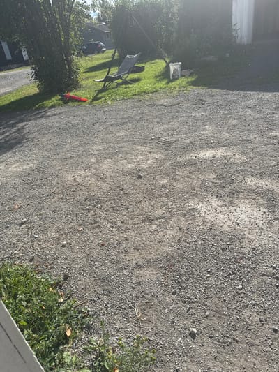The image depicts a gravel driveway or yard in Moelv, Norway. The foreground is dominated by the rough texture of the gravel. In the mid-ground, there is a patch of green grass with some vegetation. Among the grass, a red and blue toy or water gun lies on the ground. Further back, a gray lounge chair is set up on the grass, and behind it, a dark-colored SUV is parked on a paved surface, suggesting the presence of a residential area or a parking lot adjacent to a building. A white structure, possibly a house or shed, is visible to the right. Lush green trees and bushes are present throughout the scene, casting shadows and contributing to the outdoor ambiance. The sky is bright, indicating daylight.