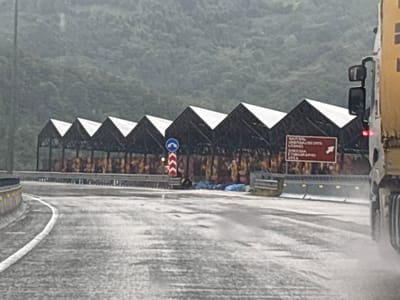 The image depicts a rainy day scene in Ubisa, Georgia, near a roadside structure with a series of triangular roofs. The foreground shows a wet road with a white line marking the lane. In the middle ground, a large red sign with white text, partially obscured by the rain and glares, points to the right, indicating a direction. A blue and white circular sign with directional arrows is also visible near the sign. Behind the sign, a long structure with multiple peaked roofs stretches across the landscape, appearing to be some form of covered parking or storage. The background is dominated by a lush, green, and densely forested mountainside. To the far right, the back of a yellow truck is partially visible, its rear lights illuminated. A person, appearing to be sitting or lying down, is visible near the base of the sign structure, partially covered by a blue tarp. The overall atmosphere is damp and overcast, with visible rain on the road and a general sense of subdued activity.