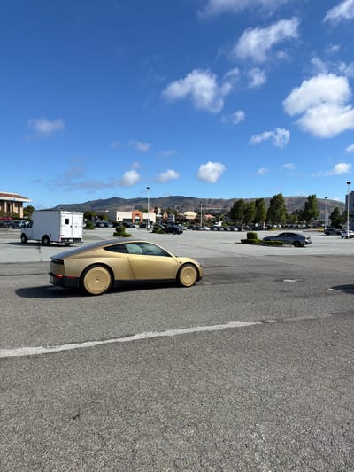 This media file captures an outdoor scene in a parking lot, likely in San Bruno, USA, given the context. The primary focus is a futuristic-looking gold car with unique, disc-like wheel covers, parked in the foreground. Behind the gold car, several rows of other vehicles are visible, suggesting a dealership or a large parking area. In the middle ground, there are trees and buildings, possibly part of a commercial area or shopping center. A white box truck is also present. The background features rolling hills or mountains under a clear blue sky dotted with fluffy white clouds. The lighting suggests it is daytime, likely a sunny afternoon. There is no visible text in this media file. The overall impression is that of a bright, possibly warm day in a suburban or commercial setting with an eye-catching vehicle.