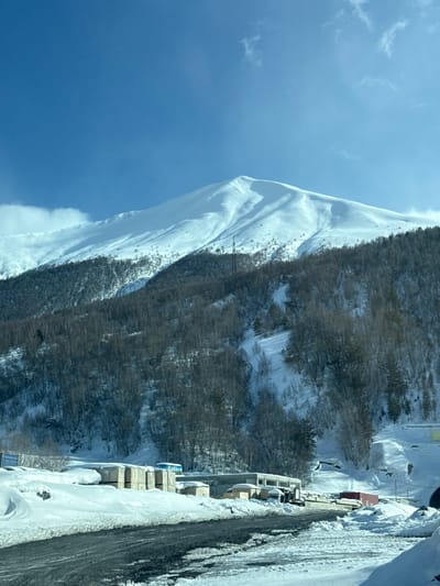 The scene depicts a winter mountainous landscape in Almasiani, Georgia, under a clear blue sky with minimal cloud cover. A prominent snow-covered mountain peak dominates the background, with its lower slopes transitioning into a dense forest of bare deciduous trees and some evergreens, also covered in snow. A dark metal lattice tower is visible on the wooded mountainside, extending towards the snowline. In the midground, several low-rise structures, consistent with industrial or storage buildings, are partially obscured by snow, alongside stacks of what appear to be building materials. A paved road surface, exhibiting patches of snow and ice, occupies the foreground. A dark vehicle and a red cargo container are identifiable near the buildings. No individuals are present.