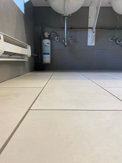 The image displays an indoor utility area, likely a public restroom or kitchen, located in Lawrence Township, United States. The floor consists of light-colored, rectangular ceramic tiles with dark grout lines. The lower walls are clad in dark gray rectangular tiles. Overhead, the undersides of two white porcelain sinks are visible, along with their chrome P-traps and exposed copper supply lines connected to chrome wall-mounted faucet valves. A white metal support bracket is affixed to the wall under the right sink. A white and blue 3M HF-PGF water filter cartridge, inscribed with the date "5-9-2023", is mounted on the wall via copper piping. To the left, a white baseboard heater or ventilation unit is attached to the wall. A black rubber plunger rests upright on the floor in the corner, positioned behind the water filter. No individuals are present, and no active events are depicted.