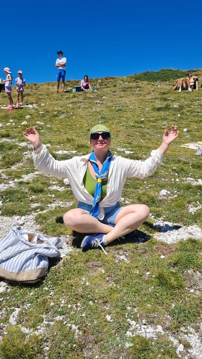 A woman is sitting cross-legged on a grassy slope, outdoors, with her arms outstretched and palms facing upwards. She is wearing a green tank top, a white unbuttoned shirt, denim shorts, blue sneakers, sunglasses, and a green bandana. A blue and yellow scarf is tied around her neck. Next to her on the left is a striped tote bag. In the background, on the same slope, other people are scattered across the grass, some sitting, some lying down, under a clear blue sky. The terrain appears to be rocky and uneven. The scene suggests a recreational outing in a mountainous or hilly natural environment. The specific location within L'Aquila, Italy, is not identifiable from the image.