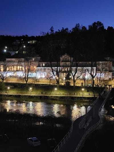Night scene depicts a building with multiple floors illuminated with lights. A river reflects these lights, creating a shimmering effect. A bridge extends over the river, connecting both sides. Bare trees are aligned along the river bank, interspersed with lampposts. The sky is a deep blue, transitioning into darkness towards the top. A hillside covered with trees forms the backdrop.