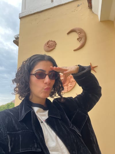 A portrait-oriented photograph, rotated 90 degrees counter-clockwise, features a person with dark, curly hair and sunglasses, looking directly at the camera with a slight pout. They are wearing a black velvet jacket over a white t-shirt, on which faint, dark lettering is visible, including what appears to be the letter "N". One hand is raised to their forehead, with fingers slightly spread, and a gold bracelet is visible on their wrist.

The background consists of a textured, light yellow-beige wall adorned with three decorative, terracotta-like objects: a crescent moon with a face, a round sun with a face, and a starfish. The wall shows some signs of wear and minor cracking. Below the wall, an overcast, cloudy sky is visible, along with glimpses of green trees in the distant background. The scene suggests an outdoor setting during the daytime, possibly in a location like Èze, France, given the architectural and decorative cues. The overall mood is casual and posed.