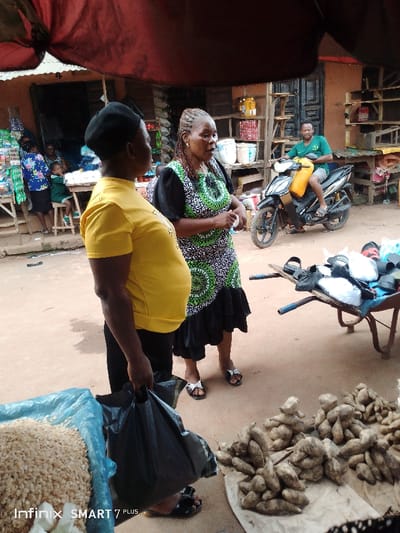 Two women converse in a bustling outdoor market, one in a yellow shirt and the other in a patterned dress. A motorcycle rider waits nearby. A wheelbarrow holds items, and various goods such as yams and grains are displayed for sale. Market stalls are visible in the background, adding to the lively atmosphere.