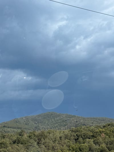 This media file depicts a scene from Dolno Varshilo, Bulgaria. The primary subjects are the sky, a forested mountainside, and what appears to be a reflection or lens flare. The sky is a pale blue with scattered clouds, suggesting daytime. There is a thin, dark line running vertically up the left side of the frame, possibly a cable or wire.

Two large, blurry, oval-shaped objects are visible in the sky, which could be reflections from a window or lens artifacts. In the middle-right portion of the frame, the side of a densely forested mountain is visible, covered in green foliage. The trees appear to be evergreen.

There is a small, dark speck in the sky near the mountain, which might be a bird or a distant object. The overall lighting indicates it is daytime, likely mid-morning or afternoon, given the brightness of the sky. The weather appears clear with some cloud cover. There is no discernible text in the image. The scene evokes a sense of being outdoors, possibly in a mountainous or elevated area with a view of the natural landscape. The image has a slightly tilted orientation, with the mountainside running diagonally from the bottom left to the top right of the frame.