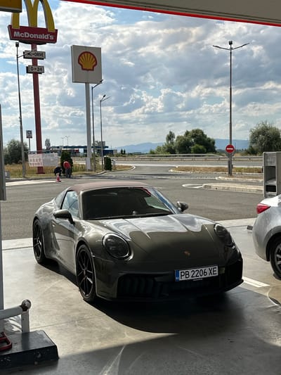 The image shows a dark grey Porsche convertible parked under a covered area at a McDonald's and Shell gas station complex. The McDonald's sign is visible in the upper left corner, along with a Shell logo. The car has a Bulgarian license plate that reads "PB 2206 XE". In the background, there are roads, trees, and mountains. Two people are visible near a red traffic cone, one wearing a red shirt and the other in dark clothing. The lighting suggests it is daytime. The location is identified as Drazhevo, Bulgaria.
