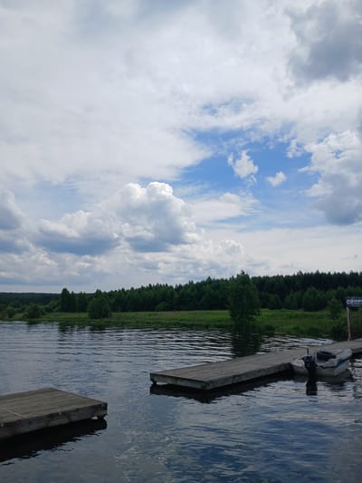 This image shows a body of water with a wooden dock extending into it. A small boat is moored to the dock. In the background, there is a lush green forest and a cloudy sky. The scene appears to be a natural landscape, likely a lake or river, given the presence of the dock and boat. There is a sign on the right side of the dock that appears to have Cyrillic writing on it, indicating it is likely located in Russia. The overall impression is one of tranquility and natural beauty.