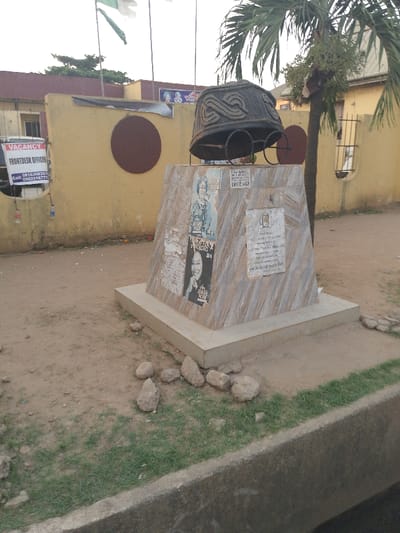 An outdoor scene shows a monument with a large hat and glasses atop a marble-like base. The base is adorned with posters and plaques. Several rocks are scattered around its base. Behind the monument is a yellow wall with a dark circle on it and a "Vacancy" sign hanging to the left. A palm tree stands to the right, partly obscuring a building in the background. Nigerian flags are displayed on poles in the distance.