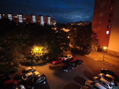 A nighttime, elevated view captures a residential area with apartment buildings and parked cars. The sky is a dark blue with some clouds. In the background, several multi-story apartment buildings with illuminated windows are visible, suggesting a high-density living area. In the foreground and mid-ground, a paved parking lot is filled with various cars of different colors and models. Some cars are parked in diagonal and parallel spots. Trees with dense foliage are scattered throughout the scene, some illuminated by streetlights, casting shadows on the pavement. A prominent streetlight with a bright yellow glow is located in the mid-ground, illuminating the surrounding trees and a portion of the parking lot. A large, orange-toned building is on the right side of the frame, with some windows lit from within. The overall atmosphere is nocturnal and urban, with artificial lighting playing a significant role in illuminating the scene. The location is identified as a residential area in the city of Most, Czechia.