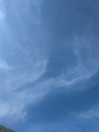The image shows a daytime sky over Chunkurchak, Kyrgyzstan. The sky is mostly blue with wispy white clouds spread throughout. A hint of green foliage appears in the bottom right corner of the frame. There are no people, animals, or other discernible objects in the main portion of the view. The scene suggests a clear, possibly high-altitude environment.