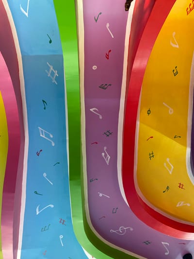This is a close-up, low-angle shot of a colorful, curved ceiling decorated with painted musical notes. The ceiling is divided into several vertical stripes of bright, solid colors: pink, light blue, green, purple, red, and yellow. White lines outline the edges of these color stripes. On each colored stripe, various musical notes and symbols are painted in white, red, and green. The lighting suggests it is daytime, with shadows casting across the yellow section. The image was captured in Bang Sare, Thailand.
