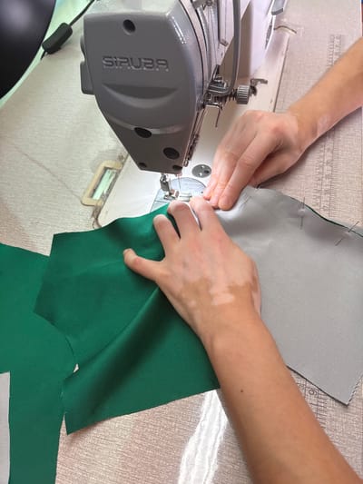 The image shows a close-up, high-angle view of a person's hands operating a "SIRUBA" brand sewing machine. The machine is positioned on a table with a textured surface. Two pieces of fabric are being worked on: one is a vibrant green, and the other is a light gray. The green fabric is to the left and is partially under the presser foot of the sewing machine. The gray fabric is to the right, pinned to the green fabric along an edge. The person's hands, which exhibit patches of vitiligo, are carefully guiding the fabric through the sewing machine. A ruler is visible on the table to the right of the fabrics, marked with measurements in centimeters. The background is blurred, but appears to be an indoor setting, possibly a workshop or studio. There is no indication of the city of Ilion, Greece, in the image or its surrounding context.