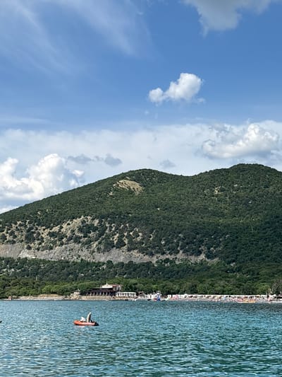 This image captures a coastal landscape in Bol'shoi Utrish, Russia, under a clear blue sky with scattered white cumulus clouds. The foreground is dominated by a body of calm, turquoise water, exhibiting gentle ripples across its surface. In the mid-ground, a small, orange inflatable boat with two individuals on board floats on the water; one person is standing towards the stern, while the other is seated.

Beyond the water, the shoreline features a mix of natural and man-made elements. To the left, a multi-story building complex with reddish-brown tiled roofs and white facades is visible, nestled among green trees, suggesting a resort or restaurant. Extending further to the right along the coast, a populated beach area is identifiable by numerous white beach umbrellas and sun loungers arranged in rows. Several people are present on the beach. A dense band of lush green trees forms a natural barrier behind the beach and structures, extending to the base of the mountains.

The background is dominated by a large, forested mountain covered in dark green trees and scrub. A distinct horizontal geological feature, possibly an exposed rock or eroded earth band, cuts across the lower to mid-slope of the mountain. The mountain peaks rise gently against the horizon.