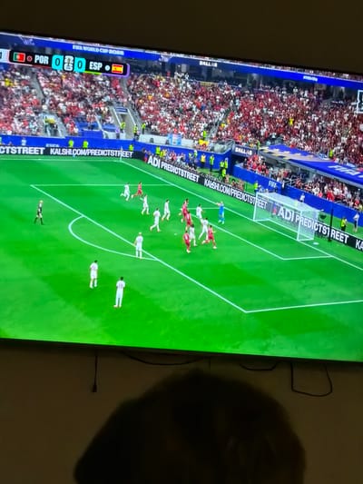 The image shows a television screen displaying a soccer match. The scoreline indicates Portugal (POR) is playing Spain (ESP) with a score of 0-0. The match is part of the FIFA World Cup 2023. The field is green with white markings. Several players in white and red uniforms are visible on the field, engaged in the game. Spectators are in the stands in the background, many wearing red. Advertising banners are displayed along the side of the field. The photograph is taken from an angle, with a dark shadow of a person or object obscuring the bottom portion of the screen and the wall below it. There are cables hanging from the bottom edge of the television. The image does not provide any specific information about the location within the city of El Escorial, Spain.