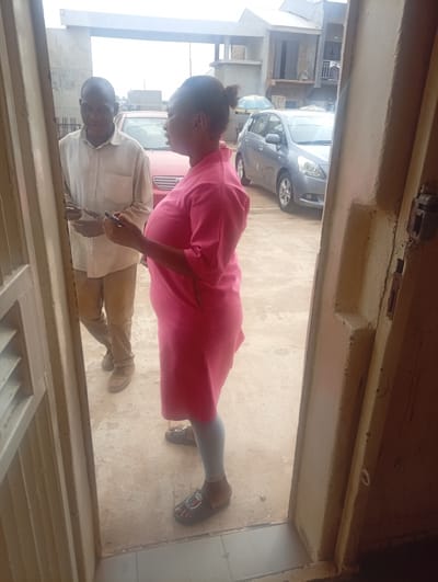In Kinkinau, Nigeria, a man and a woman are engaged in a conversation. The man, wearing a light-colored long-sleeved shirt and brown trousers, stands on the left, holding a smartphone in his left hand. He appears to be smiling, looking towards the woman. The woman, on the right, is dressed in a bright pink, knee-length dress and light gray leggings. She is also holding a smartphone and facing the man. Her expression is neutral, but her posture suggests engagement in the conversation.
They are standing outdoors, possibly in front of a building or entrance, with a beige-colored gate or door frame visible in the foreground on the left. In the background, two cars are parked: a red car on the left and a silver hatchback on the right. Beyond the cars, buildings with multiple stories and balconies are visible, suggesting an urban or semi-urban setting. The sky is overcast, indicating a cloudy day. The overall scene appears to be a casual interaction, likely during daylight hours. There is no visible text in the image.
