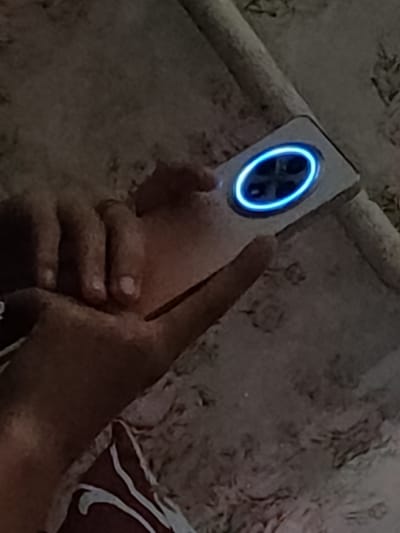 A hand is holding a white smartphone with a circular camera module that has a blue illuminated ring. The phone is positioned on a textured surface that appears to be a blanket or carpet. The background is dark and indistinct. The image does not provide any information about the city of Ternovka, Moldova.