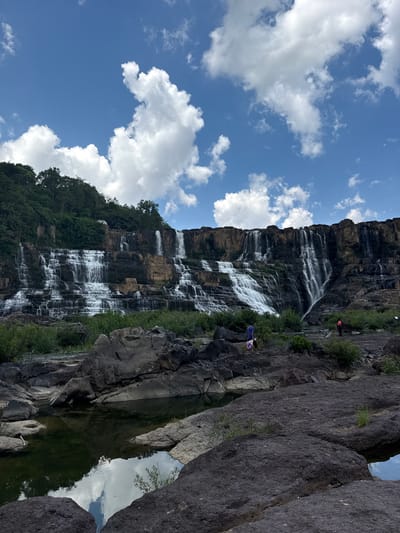 This image depicts the Pongour Waterfall, a multi-tiered waterfall located in the Tân Phú area of Vietnam. The scene is dominated by the cascading water flowing down a wide, rocky cliff face. Lush green vegetation covers the top of the cliff and borders the falls. In the foreground, dark, rugged rocks form the edge of a shallow pool of water that reflects the sky and clouds. Two individuals are visible near the base of the waterfall. One person, wearing a blue shirt and carrying a bag, is walking on the rocks. Another person, wearing an orange shirt, is standing in the distance, seemingly observing the waterfall. The sky above is a vibrant blue, dotted with fluffy white clouds.