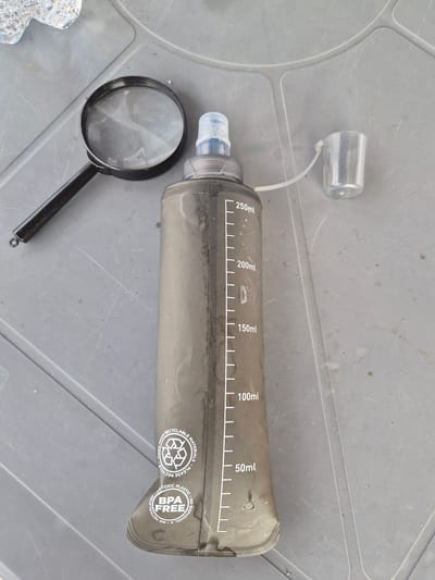 A soft-sided, black water bottle with volume markings in white is laid out on a gray, textured surface, possibly a table or plastic container lid. The bottle has a clear plastic cap attached by a thin white cord. A black-handled magnifying glass rests to the left of the bottle, and a small piece of clear, textured material is partially visible in the bottom left corner. The water bottle features markings indicating volumes of 50ml, 100ml, 150ml, 200ml, and 250ml. Circular text and symbols are printed on the right side of the bottle, including "RECYCLABLE MATERIALS," "PLEASE RECYCLE," "MADE FROM RECYCLED," "NON-TOXIC PLASTIC," "BPA FREE," and "NO BISPHENOL A. NO PHTHALATES." The scene appears to be outdoors, possibly in daylight, given the lighting and the texture of the surface. The overall impression is one of an outdoor or camping setting, with an emphasis on practical, reusable items.