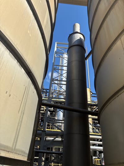 The image captures an industrial scene at a facility in Rąbczyn, Poland. Two large, cylindrical, light-colored storage tanks dominate the foreground, framing a central metallic smokestack and a complex network of industrial piping and structures. The smokestack is dark gray and appears to be made of segmented metal sections. Behind the smokestack, a white metal scaffolding-like structure is visible, along with various silver pipes and yellow railings. The sky above is a clear, bright blue with a few wispy white clouds. The overall impression is one of an active, operational industrial site under a sunny sky. There are no people visible in the image.