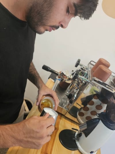 A barista in a black shirt pours milk from a silver pitcher into a small, round coffee cup. He stands in front of a shiny espresso machine and a white coffee grinder with brown polka dots, both resting on a light wooden surface. The man has dark hair and a beard. His tattooed arm is visible as he expertly creates latte art.