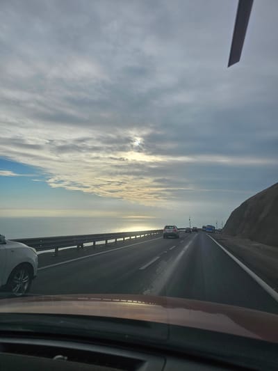 The image shows a view from a vehicle driving on a highway. The highway runs alongside the Pacific Ocean, with a rocky hillside to the right. The sky is overcast with streaks of clouds, and the sun is visible through a break in the clouds, reflecting on the ocean. There are several cars and a blue bus on the highway. The photo appears to be taken in the city of Alto Hospicio, Chile, known for its coastal highways and arid landscape.