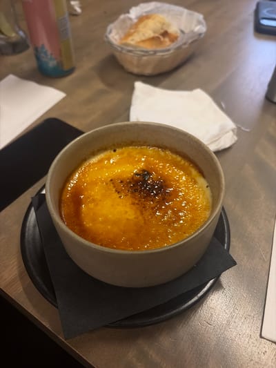 A close-up shot captures a crème brûlée in a light beige bowl, presented on a dark plate with a black napkin. The crème brûlée has a perfectly caramelized, slightly browned sugar crust, with a few darker charred spots. The custard beneath appears a vibrant yellow. The bowl is situated on a wooden table, with a blurred background showing a white napkin, a basket with bread, and a pink and yellow patterned bottle. The scene is lit with natural light, creating a warm and inviting ambiance, likely within a restaurant setting in Longueuil, Canada. There are no people visible in the image.