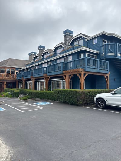 A blue building with multiple floors and balconies stands next to a parking lot. The structure features wooden support beams and stone chimneys. A white car is parked in front of the building, separated by a green hedge. Parking spaces are marked on the asphalt, including a handicapped spot. The sky is overcast.