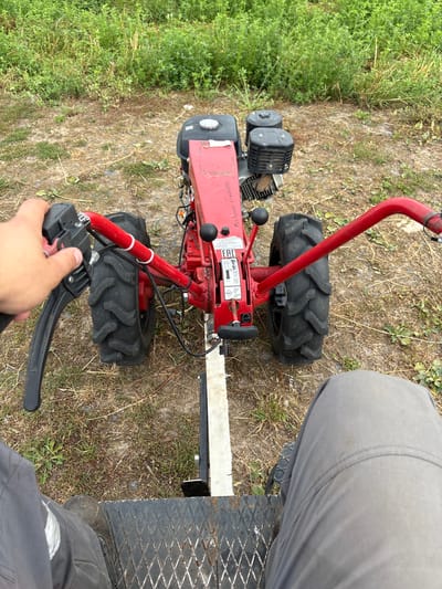 The image shows a person's legs and hand operating a red walk-behind tractor. The tractor has two large black tires and a red frame. The person is wearing gray pants and a dark boot on their left foot. The tractor is on a dirt path surrounded by green vegetation and dry grass. The background shows more greenery and an overcast sky. The scene appears to be outdoors in a rural area, possibly a field or a garden. No specific landmarks or structures are visible that would indicate a precise location within Урыцкі сельскі Савет, Belarus.