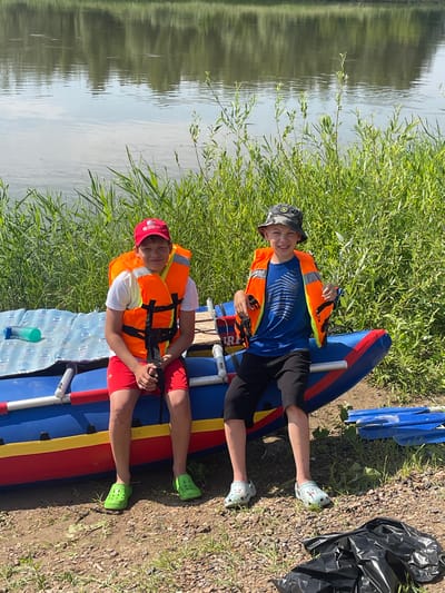 Two young boys are sitting on the edge of an inflatable raft on the shore of a river. The boy on the left is wearing a white t-shirt, an orange life jacket, red shorts, and green clogs. He has a red baseball cap on. The boy on the right is wearing a blue t-shirt, an orange life jacket, black shorts, and light blue clogs. He has a camouflage bucket hat on. The raft is blue with red and yellow accents. Behind the raft, tall green grass and reeds grow along the riverbank. The river itself is calm and reflects the trees and sky on the opposite bank. The ground in front of the raft is covered in dirt and small stones. A black trash bag is visible on the ground to the right of the raft. The overall setting appears to be a natural, outdoor environment, possibly a park or recreational area near a body of water. The image was taken in Nezhinka, Russia.