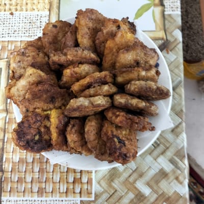 A plate of fried food, likely cutlets or patties, is presented on a textured placemat with a woven bamboo pattern. The food is golden brown and appears to be slightly greasy, with some pieces showing a darker, crispier exterior. The placemat is light brown and beige, with a repeating basketweave design. A white napkin is visible underneath the plate. The background is blurred, but suggests an indoor setting with a light-colored surface and some greenery from a plant. The scene is likely a casual meal or snack being served.