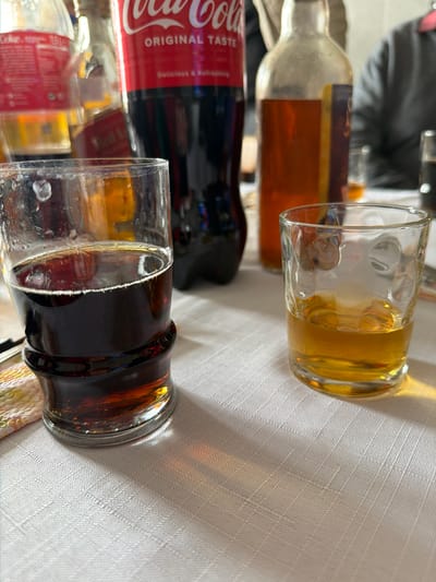 The image shows a table setting with two glasses, a bottle of Coca-Cola, and another bottle of an amber-colored liquid, possibly whiskey or rum. One glass contains a dark brown beverage, likely Coca-Cola, while the other contains a yellow liquid, possibly the amber-colored liquor. The table is covered with a textured white tablecloth. The scene appears to be indoors. The image does not provide enough information to confirm it was taken in Dulevo, Bulgaria.