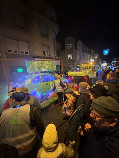 The scene depicts a nighttime street event in Wasserbillig, Luxembourg. A crowd of individuals observes a procession along a city street flanked by multi-story buildings. In the left foreground, a flatbed trailer carries a green Ford Ka automobile, marked with the number "58" on its side. The vehicle and trailer are decorated with multi-colored string lights and surrounded by hay bales. Above this float, a banner displays text in German: "AUTOCROSS AUSDAUER-RENNEN IN HELLANGE" and the date "18. MAI 2020," along with an email address and "FB: CRAZY FRIENDS." Several people are positioned near this float, including an individual in a light-colored reflective vest and another in the lower-left foreground wearing a reflective vest emblazoned with "CRAZY FRIENDS."

Further down the street to the right, a yellow utility vehicle, possibly a customized float, also adorned with string lights and an orange beacon, is visible as part of the procession. Spectators, dressed in winter attire including hats and coats, line both sides of the street, with a young child in a patterned jacket observed near the green car float. Buildings on either side of the street feature illuminated windows, some with rolled-down shutters. A blue pedestrian crossing sign is visible on the right side of the street. The environment is illuminated by streetlights, building lights, and the decorative lights of the parade elements.