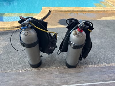 Two scuba diving tanks with their regulators and backpacks are standing upright on a textured gray surface next to a swimming pool. The tanks are silver and appear to be made of aluminum. The regulators have black and silver components, and one is attached to a blue strap. Yellow hoses are visible connecting to the regulators. The backpacks are black and appear to be made of nylon.
The swimming pool is to the left and behind the tanks, filled with clear blue water. The edge of the pool is made of light-colored stone. The ground surface is a mix of small gray pebbles and some wooden planks.
The lighting suggests it is daytime, likely morning or late afternoon, with distinct shadows cast on the ground. The weather appears to be clear and sunny. The presence of scuba gear and a swimming pool suggests a recreational or training context, possibly at a dive resort or hotel. The overall scene is peaceful and ready for activity.
Visible text includes "CRESSI" on one of the black backpacks. There are also markings on the silver tanks, but they are not clearly legible in the image. The location context is Kubu, Indonesia, which is known for its diving sites.