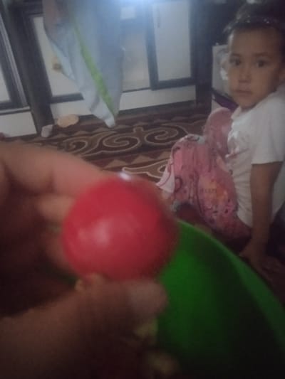 In the city of Кызыл-Булак, Kyrgyzstan, a child is seated on a patterned rug, wearing a white t-shirt and pink pants with a floral pattern. To the left of the child, a hand is visible holding a red, spherical object, possibly a toy, which is partially obscuring a green object below it. In the background, a curtain with a green stripe is visible, and a blurred window frame is present. The lighting in the scene appears to be artificial and somewhat dim.