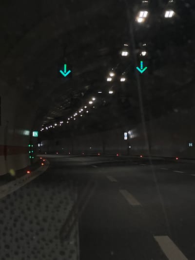 The image shows the interior of a tunnel in or near Pelev Brijeg, Montenegro. The tunnel's interior is dimly lit by overhead lights, with rows of small lights along the tunnel walls. There is a red line on the ground along with orange traffic cones. There are no people visible. The image has 2 light-green right arrows.