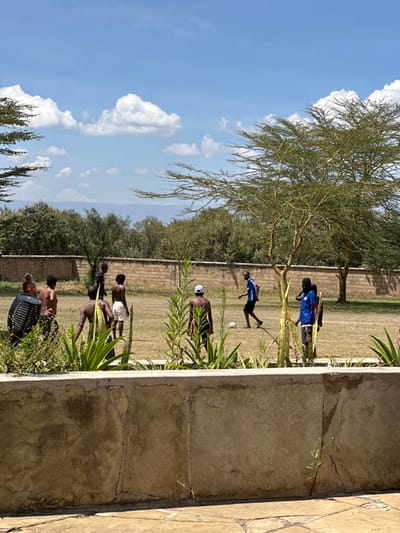 The image captures an outdoor scene in Olkaria ward, Kenya, under a bright blue sky with scattered white cumulus clouds. In the immediate foreground, a light-brown, textured concrete or stone planter box, topped with various green leafy and succulent plants, partially obstructs the view. Beyond this, a dry, sparsely green grassy field serves as the setting for approximately eight males, appearing to be young adults or teenagers, engaged in a casual game of soccer.
One individual, positioned in the right-midground, wears a blue long-sleeved shirt and dark shorts, actively kicking a white soccer ball. Another male, further to the right, wears a blue short-sleeved shirt and dark shorts. The remaining individuals are mostly shirtless, wearing dark shorts, with one individual in the left-midground wearing a white cap and another on the far left near a black plastic crate or bin. Several acacia-like trees with light green, sparse foliage are distributed across the field. A low, light-brown, block-built wall extends horizontally across the mid-background, behind which lies a denser cluster of green trees and shrubs. Distant, hazy blue mountains are visible on the horizon.