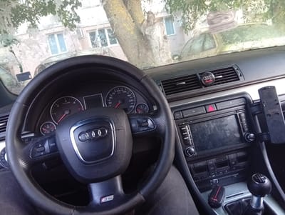 The image provides an interior view of an Audi car, likely a model from the mid-2000s based on the dashboard design. The perspective is from the driver's seat, focusing on the steering wheel, dashboard, and center console.
The steering wheel is black leather with red stitching visible on the lower spoke, featuring the Audi rings logo in the center. Buttons for controlling various functions are integrated into the wheel. The instrument cluster displays speed in km/h and RPM, with a prominent tachometer showing around 750 RPM.
The center console includes the car's infotainment system with a screen displaying what appears to be a blank or off screen, along with physical buttons for navigation, radio, and other settings. Below the infotainment system are climate control buttons, some labeled "AUTO" and "ECON." A manual gear shifter is visible, suggesting a manual transmission. An Audi key fob, also with the characteristic rings, rests near the gear shifter. A mobile phone mount is attached to the right side of the center console.
On the dashboard, an Audi air freshener is clipped to the air vent, featuring the brand's logo. Outside the car, through the windshield, a tree with rough bark is prominent, and behind it, buildings with multiple windows are visible. The windshield appears somewhat dirty or streaked.
The scene suggests the car is parked, possibly on a street in Aidemir, Bulgaria, given the context. The lighting indicates it's daytime, but the overcast conditions or the cleanliness of the windshield make it difficult to determine the exact time or weather conditions definitively. No people are visible in the frame, but the presence of the steering wheel and gear shifter implies a driver's seat. The overall impression is a typical view from inside a parked, well-maintained Audi car.