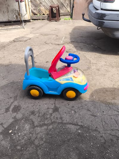 A blue and pink plastic ride-on toy car for children is parked on a cracked asphalt surface. The car has a yellow steering wheel and yellow wheels with black tires. A gray handle extends from the back of the car, suggesting it can be pushed. A small, clear plastic dome covers the front of the car, and a doll or toy is visible inside. The car has "SPACE CLUB" written on its side. In the background, there is a wooden fence, a brown plastic stool, and the rear bumper of a white car with a tow hitch. The scene appears to be in an outdoor, possibly a courtyard or parking area, with some scattered debris on the ground. The lighting suggests it is daytime. There is no indication of the specific location within Barysaw, Belarus, beyond being an outdoor urban or suburban area.