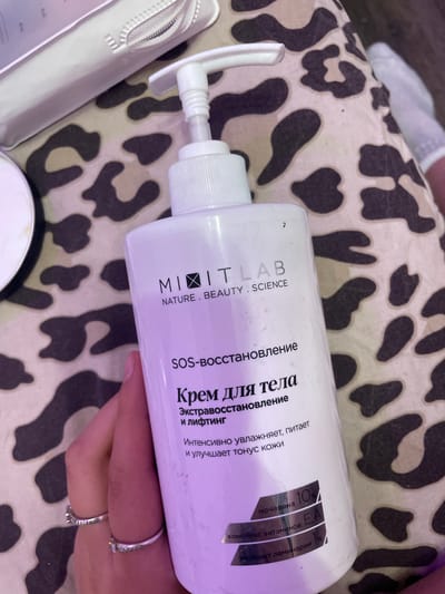 A hand with rings holds a white pump bottle of "MITIT LAB SOS-restoration body cream" on a leopard-print fabric. The bottle has Russian text indicating its purpose and ingredients. The background is a blurry close-up of the leopard print fabric and a portion of another white object, possibly a cosmetic bag. There is no visible indication of the city of Лесное Снт, Russia within the image.