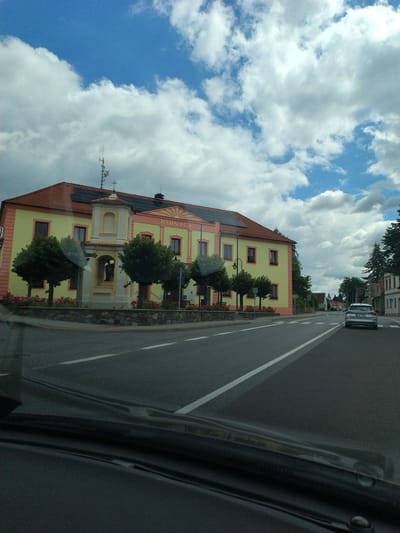 The image captures a view from inside a vehicle, looking out at a building with the word "RADNICE" visible on its facade, suggesting it is a town hall or administrative building. The building is light yellow with a red roof and decorative orange trim. In front of the building, there is a small monument with a statue and a cross. Trees and flower beds are situated between the monument and the road. A silver car is driving away on the right side of the road. The scene is set against a blue sky with white, puffy clouds. The location is identified as Bernartice, Czechia. The image depicts a street scene in a town.