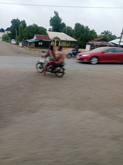 The image captures a street scene in Kwale, Nigeria, under an overcast sky. The foreground is dominated by a motion-blurred asphalt road. In the middle ground, two figures are riding a motorbike. The person in the front appears to be driving, while the person in the back is seated behind them. The motorbike is light in color. To the right of the motorbike, a red sedan is in motion. Further in the background, several buildings are visible, including houses with pitched roofs and structures with corrugated metal roofs. One building has a green-yellow facade and an arched entrance. Trees are scattered throughout the background, some dense and green, others appearing more sparse. A white van is parked on the left side of the road in the distance. A small, dark-colored structure with a red corrugated metal roof and a light blue tarp covering part of it is positioned near the road. The overall impression is one of a typical roadside environment in a Nigerian town, with ongoing traffic and residential or commercial buildings.