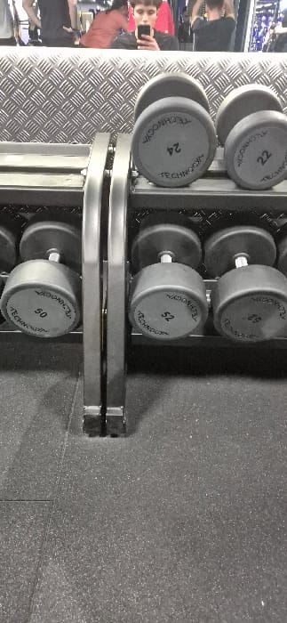 The image displays an interior view of a fitness facility located in Carnaxide, Portugal. The bottom portion of the frame shows a dark gray, speckled rubber floor. In the midground, a two-tiered metal dumbbell rack is visible, housing multiple black Technogym dumbbells with silver handles. Specific dumbbell weights clearly identifiable include 50, 52, 24, and 22, likely indicating kilograms. The background is a large mirror reflecting the gym's interior. In the reflection, a textured, light gray metallic wall is discernible behind the dumbbell rack. At least three individuals are reflected in the mirror. A young person with short dark hair, wearing a dark shirt, is centered in the reflection, actively holding a black smartphone to take a selfie. To the left of this person, another individual with longer, reddish-brown hair and a matching top is partially visible. To the right, a third person in a dark shirt is reflected with their arms raised, seemingly engaged in an overhead exercise.
