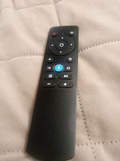 A black remote control is lying on a beige quilted surface. The remote has several buttons with icons and labels. At the top, there is a red power button. Below that, there is a circular navigation pad with a central button. Further down, there are directional arrows, a "TB" button, a microphone button that is illuminated blue, a house icon, a play/pause button, a minus button, and a plus button. The background is soft and textured, appearing to be fabric. The location is not ascertainable from the image.