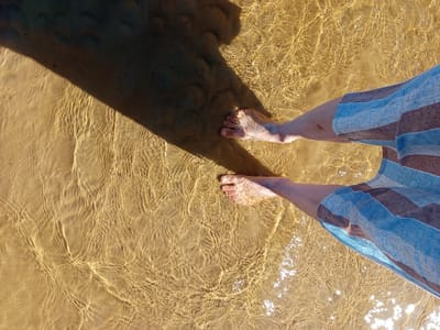 An overhead shot captures the lower legs and bare feet of an individual standing in shallow, clear water with a sandy bottom in Podborov'e, Russia. The person wears blue and brown horizontally striped shorts or swim trunks, with their calves and feet submerged in the water. Sunlight penetrates the water, creating distinct ripple patterns and bright reflections on the sandy surface beneath. A large, dark shadow extends diagonally from the upper left, partially obscuring the water and sand in the upper left quadrant of the image and falling across the area near the person's left foot. The water appears calm but shows subtle movement, creating varied light patterns.